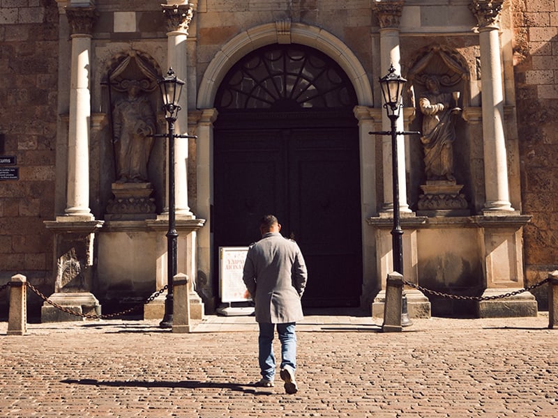 Back view of a person approaching the main entrance of a historic Spanish church. The stone facade includes columns, statues, lanterns, and a large arched doorway, with a cobblestone square in the foreground.